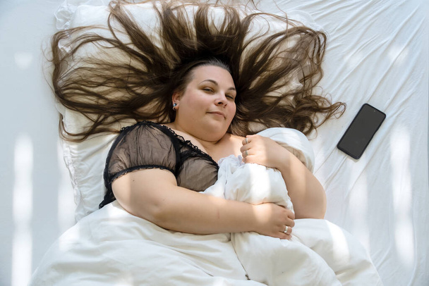 top view of happy positive beautiful cheerful fat plump cute chubby sexy plus size woman, with long hair relaxing in bed, enjoying morning sunlight stripes, smartphone, copy space - Photo, Image
