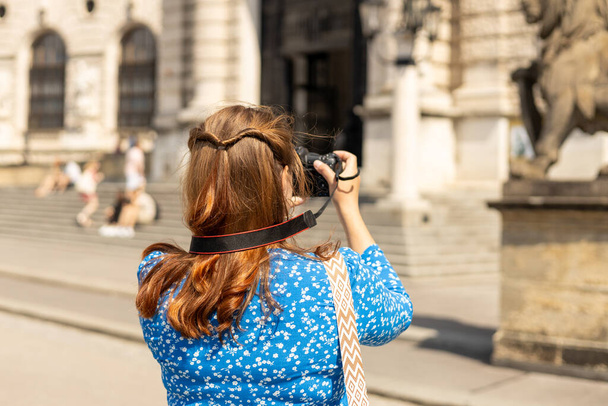 Young woman holding the camera outdoors. Beautiful hipster girl shooting with camera on city street, back view. Concept of travel, tourism and vacation in city. - Photo, Image