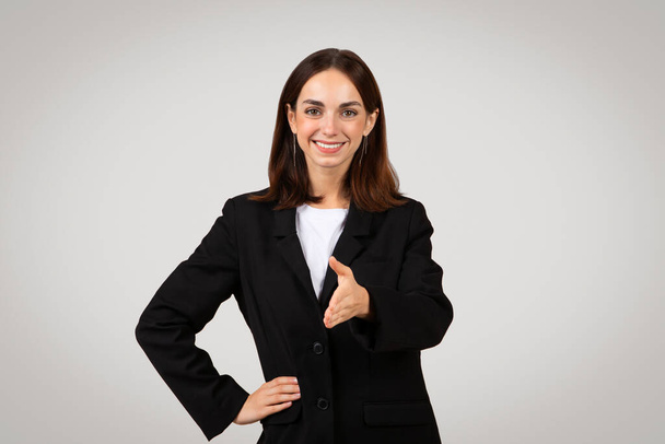 Approachable and professional caucasian young businesswoman with long hair wearing a black suit extends a handshake, suggesting a welcoming gesture or agreement, isolated on gray background, studio - Photo, Image