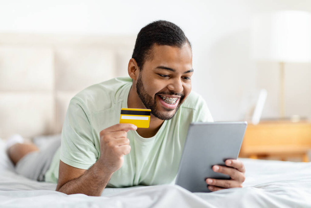 E-Commerce, Einzelhandel, virtuelles Banking. Aufgeregte junge afrikanisch-amerikanische Kerl auf dem Bett liegend, mit digitalem Tablet und Plastikbankkarte, Online-Bestellung während zu Hause bleiben am Wochenende, Kopierplatz - Foto, Bild