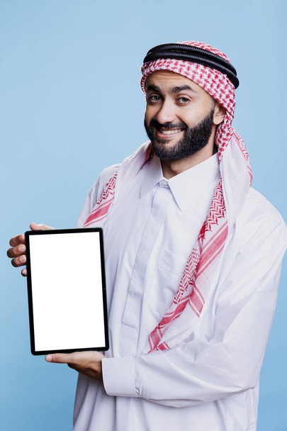 Smiling arab dressed in traditional attire holding digital tablet with white blank screen mockup and looking at camera. Muslim man promoting portable gadget empty touchscreen for application ads - Photo, Image
