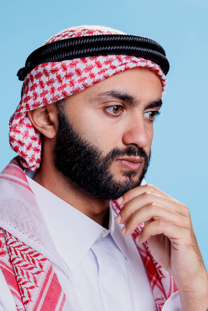 Geleneksel başlıklı Müslüman adam düşünüyor ve çözüm düşünüyor. Arap bir insan, başörtüsü takıyor, çenesini ovalıyor ve yoğun bir ifadeyle yüzünü çeviriyor. - Fotoğraf, Görsel