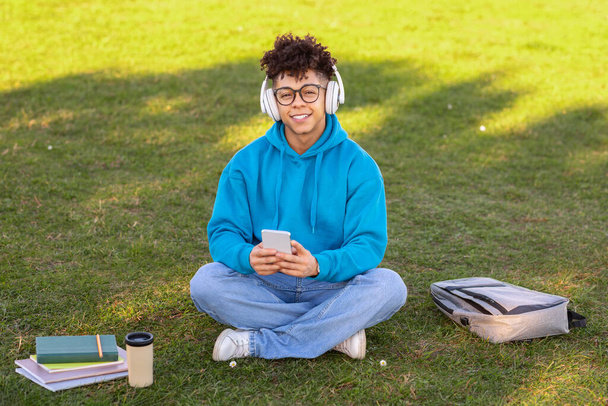 Glimlachende zwarte student kerel met een koptelefoon houdt zich bezig met mobiele telefoon, het luisteren naar muziek en educatieve podcasts tijdens het leren van outdoor, zitten met zijn rugzak buiten - Foto, afbeelding