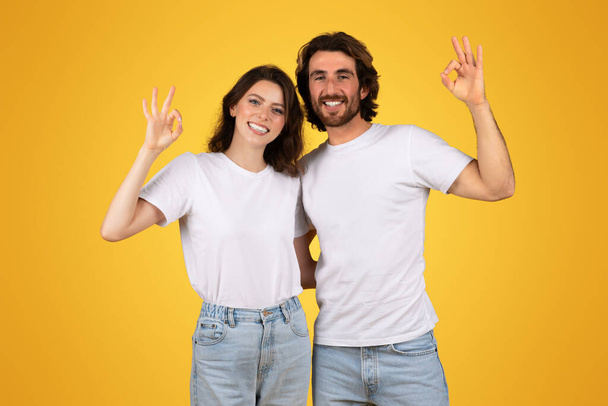 Vidám európai férfi és nő fehér pólóban és farmerban, akik az OK kézmozdulatot teszik, magabiztosan mosolyognak meleg sárga háttérrel, ami az elégedettséget és a pozitivitást szimbolizálja. - Fotó, kép