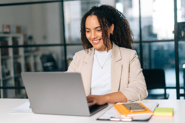 Amazing curly haired hispanic or brazilian young woman in stylish elegant clothes, office employee, small business owner, working on a laptop while sitting at her workplace in the modern office, smile - Photo, Image
