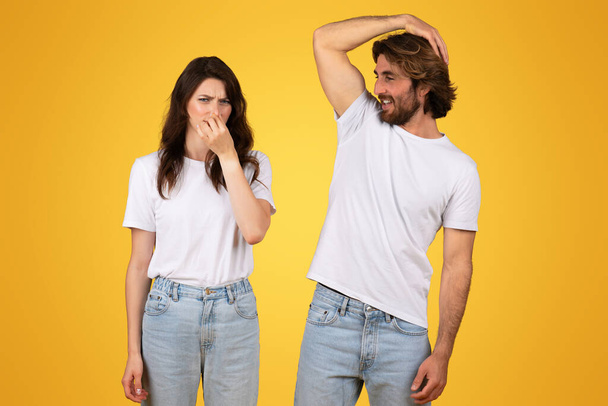 Eine Europäerin kneift sich mit angeekeltem Gesichtsausdruck die Nase, während sich ein Mann lächelnd den Kopf kratzt, beide tragen weiße T-Shirts und Jeans vor einem knallgelben Hintergrund. - Foto, Bild