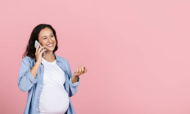 Happy young pregnant woman talking to her expecting friend on phone, gesturing and smiling, looking at copy space isolated on pink studio background, sharing pregnancy experience - Photo, Image