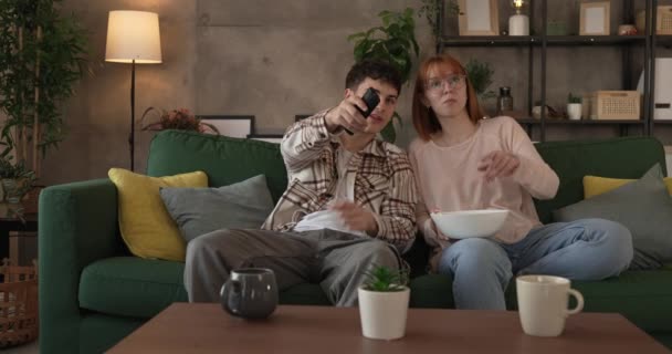 Pár kaukázusi férfi és nő ül otthon kanapén TV-t néz - Felvétel, videó