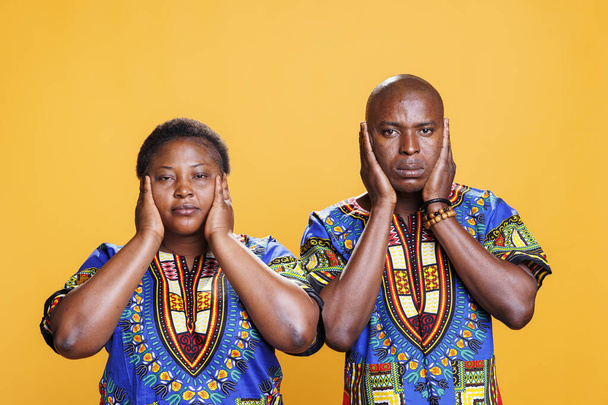 Αφρικάνικο ζευγάρι καλύπτει τα αυτιά με τα χέρια, δείχνοντας δεν ακούει κακό τρεις σοφούς πιθήκους πορτρέτο. Άντρας και γυναίκα αγνοούν τον δυνατό θόρυβο και κοιτάζουν την κάμερα στο φόντο του στούντιο - Φωτογραφία, εικόνα