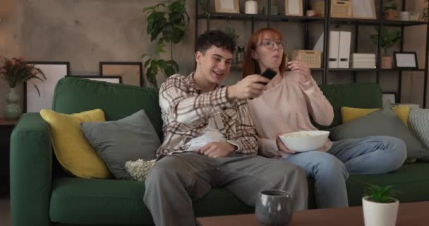 Pár kaukázusi férfi és nő ül otthon kanapén TV-t néz - Felvétel, videó