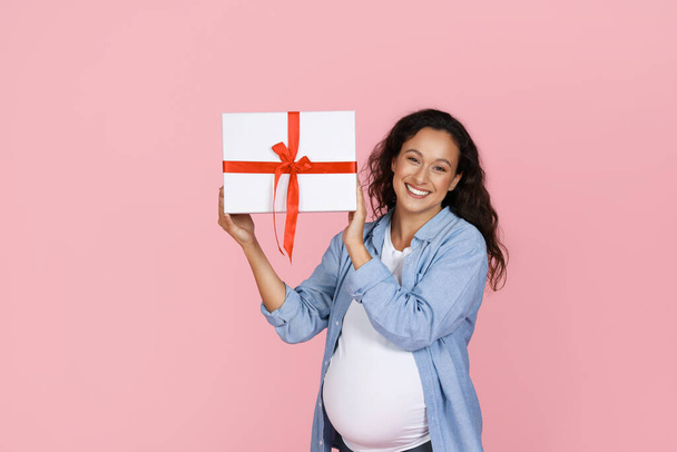 Παιδικό πάρτι. Χαρούμενη νεαρή έγκυος γυναίκα σε casual με μεγάλη κοιλιά δείχνει κουτί με δώρο, απομονώνονται σε ροζ φόντο στούντιο. Αναμονή για το παιδί, συγχαρητήρια για τις διακοπές, ιδέα για σήμερα - Φωτογραφία, εικόνα