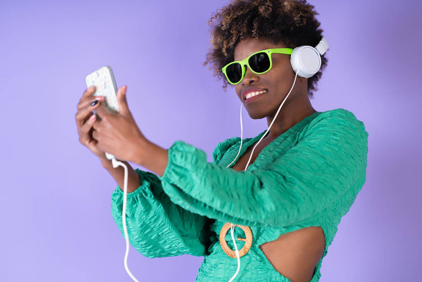 Mladá hezká černá žena s kudrnatými vlasy nosí sluneční brýle a poslouchá hudbu na svém smartphonu se sluchátky na šeříkovém pozadí, s kopírovacím prostorem. Koncept štěstí. - Fotografie, Obrázek
