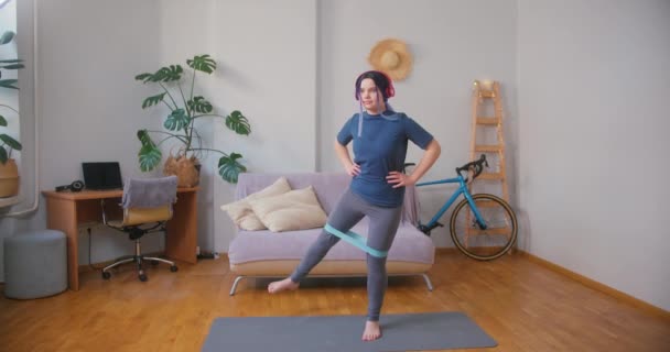 Young plus size latin hispanic teenager girl exercising with resistance rubber band at home. Cozy home interior with indoor plants. Online fitness, urban jungle concept. - Footage, Video