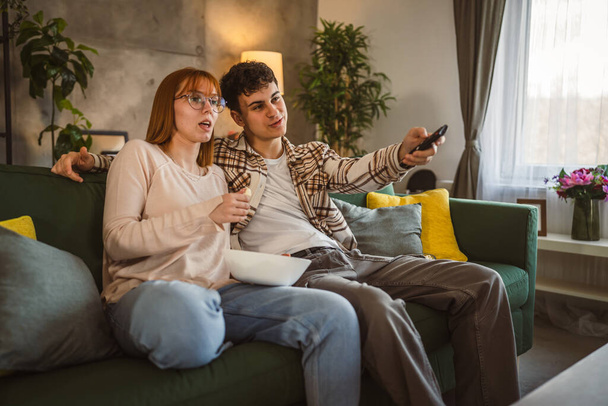 Пара белых мужчин и женщин сидят дома на диване и смотрят телевизор. - Фото, изображение