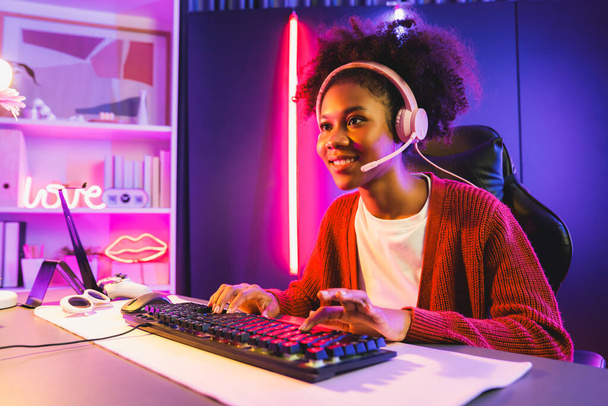 Afroamerikanische Mädchen Streamer spielen Online-Kampf mit Esport qualifizierte Team mit Kopfhörern in neonfarbener Beleuchtung Raum. Gespräche mit anderen Spielern, die Strategien planen, um Konkurrenten zu gewinnen. Geschmacksmacher. - Foto, Bild