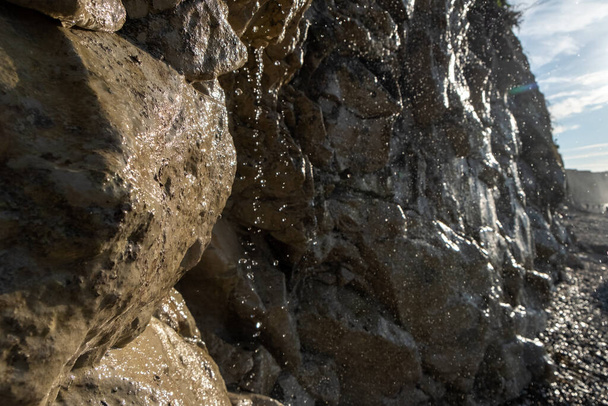 Dieses Bild zeigt eine detaillierte Nahaufnahme des Wassers, das an einem sonnigen Strand eine raue, strukturierte Felswand hinunterstürzt. Das Sonnenlicht fängt einzelne Tröpfchen ein und unterstreicht ihre Klarheit und die komplizierten Wege - Foto, Bild