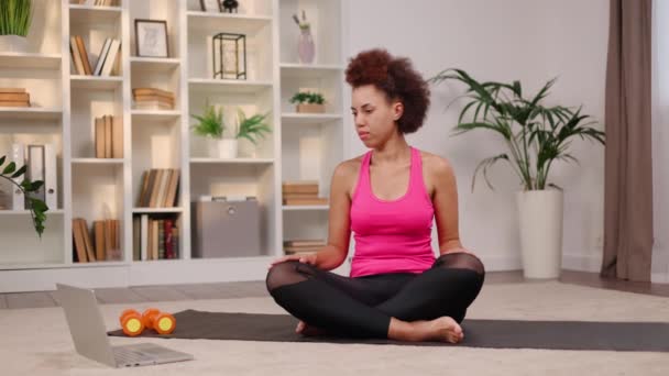 Young african american woman doing yoga with online trainer on modern laptop and repeating movements. Calm female wearing sport clothes relaxing body on black yoga mat in cozy living room. - Footage, Video