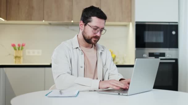 Side view of competent male freelancer working remotely on portable laptop and looking at camera at home. Handsome young caucasian man using modern device for doing successful tasks indoors. - Video, Çekim