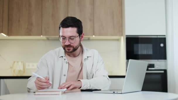Multitasking caucasian male sitting at workplace and making notes in personal notebook while typing on wireless laptop indoors. Calm smiling man using modern gadget for productive performance of task. - Кадры, видео