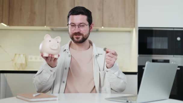 Happy unshaved entrepreneur sitting by desktop with pc with pink ceramic money box and euro in hands. Inspired businessperson receiving profit from long term investment and collecting savings. - Footage, Video