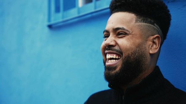 Ein fröhlicher junger schwarzer brasilianischer Mann lacht und lächelt vor einer städtischen blauen Wand, das Gesicht einer südamerikanischen Person, die sich mit Freunden aus dem Off unterhält - Foto, Bild