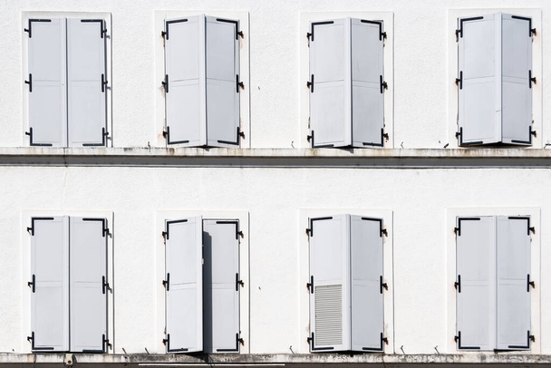 Kapalı beyaz panjurlu sekiz pencere, biri aralık, eski beyaz bir binanın ön cephesinde. - Fotoğraf, Görsel