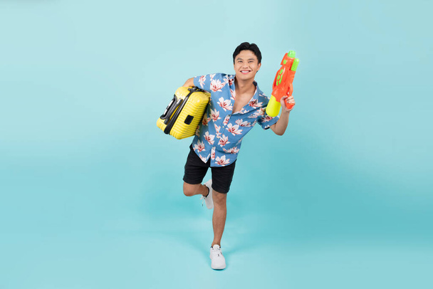 Gelukkig knappe Aziatische toerist in de zomer outfits met waterpistool en koffer in blauwe kleur studio geïsoleerde achtergrond - Foto, afbeelding