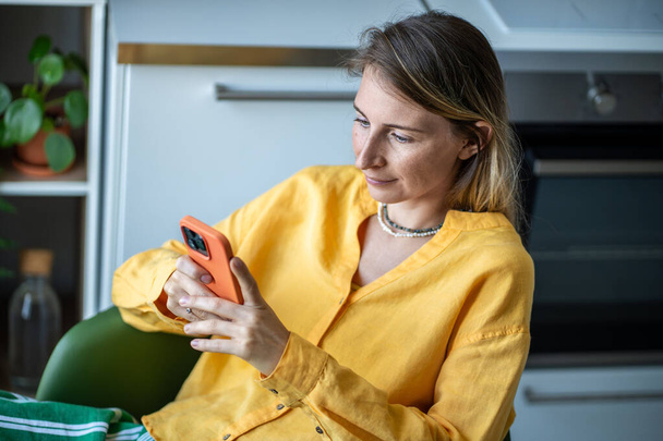 Расслабленная шведская женщина читает онлайн-сообщения, просматривая новости социальных сетей дома. Скандально известная длинноволосая женщина с помощью приложений для смартфонов сидит на стуле на кухне. Цифровая зависимость. - Фото, изображение