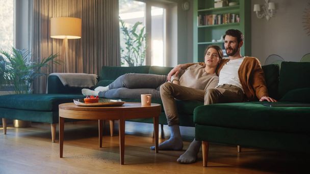 Couple Watches TV while Sitting on a Couch in the Living Room. Girlfriend and Boyfriend Lying Embracing. Spending Weekend Together at Home with Stylish Interior. - 写真・画像