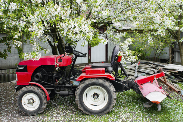 Piros kis modern traktor áll a gazdaság hátterében egy tavaszi táj. A kormányrúd a traktorhoz van erősítve. Kis mezőgazdasági gépek. Mezőgazdasági munkaterv. - Fotó, kép