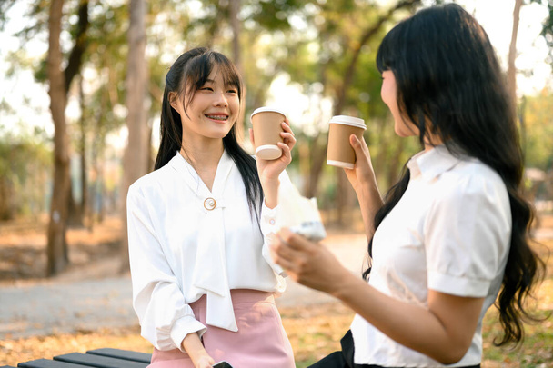 Mutlu genç Asyalı kadınlar kağıt bardaklardan kahve içip parkta sohbet ediyorlar.. - Fotoğraf, Görsel