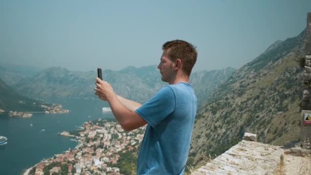 Человек-турист фотографируется на смартфоне, стоящем на горе Ловчен над Которским заливом и старым городом Черногории. Мужчина-путешественник фотографирует на мобильный телефон прекрасный вид на Адриатический фьорд летом - Кадры, видео