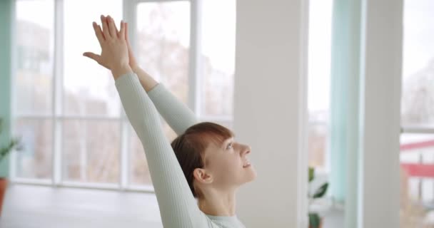 Een slanke vrouw die yoga doet in de studio. Handheld shot van barefoot vrouw in lichte sportkleding doet Crescent Lunge op de knie en Half Splits poseert tijdens yoga sessie in zonovergoten studio. - Video