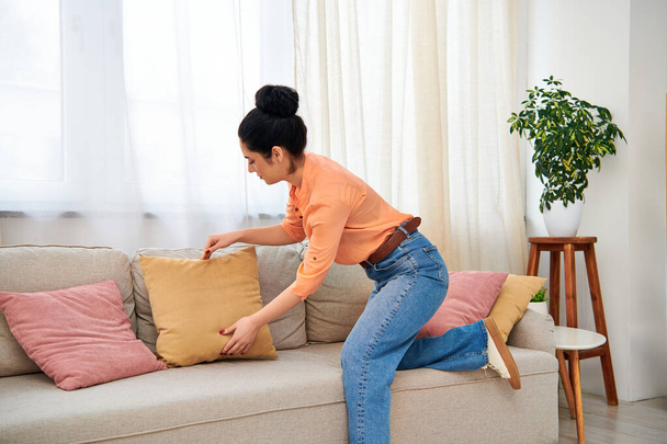 A stylish woman in casual clothes sits gracefully on a couch, clutching a cozy pillow. - Photo, Image