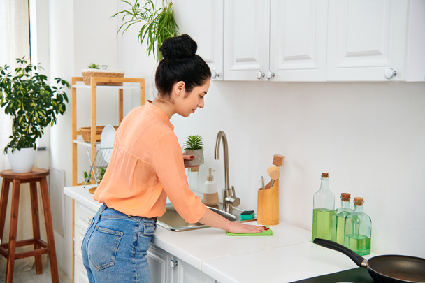 Kobieta w luźnym stroju stoi przy zlewie kuchennym, z patelnią na ladzie. Wygląda na skupioną i pogodną, gdy wykonuje swoje obowiązki domowe.. - Zdjęcie, obraz