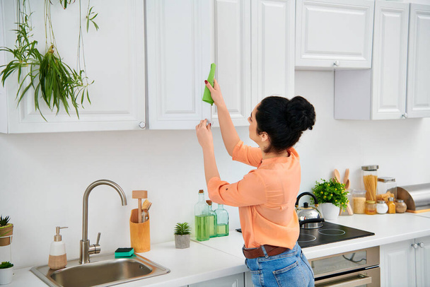 Una mujer con estilo en traje casual metódicamente limpia el fregadero de la cocina con un trapo verde vibrante, trayendo una limpieza radiante. - Foto, Imagen