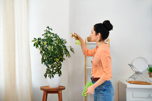 Egy narancssárga inges, elegáns nő gondosan tisztít egy növényt, szeretetet és törődést mutatva az otthoni környezetével.. - Fotó, kép