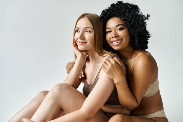 Δύο διαφορετικές γυναίκες σε άνετο παστέλ εσώρουχα κάθονται στενά σε ένα λευκό φόντο. - Φωτογραφία, εικόνα