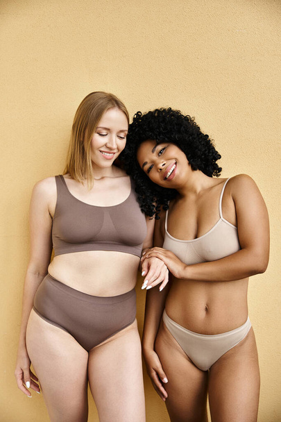 Two diverse women elegantly pose in cozy pastel underwear. - Photo, Image