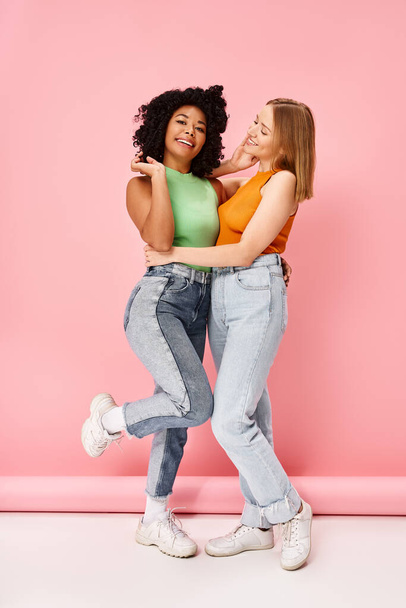 Two women in casual attire embrace, smiling against a pink backdrop. - Photo, Image