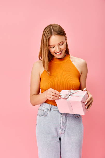 Μια γυναίκα με πορτοκαλί μπλούζα κρατά ένα ροζ κουτί δώρου, αποπνέοντας ζεστασιά και ενθουσιασμό.. - Φωτογραφία, εικόνα