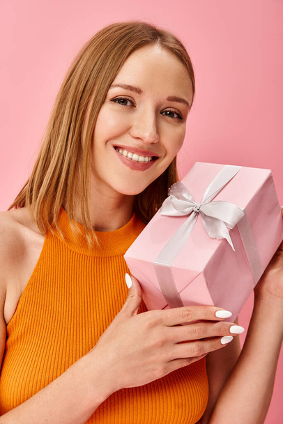 Egy nő narancssárga felsőben örömmel tart egy rózsaszín ajándékdobozt.. - Fotó, kép