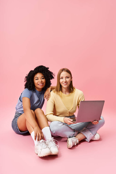 Две разные женщины в повседневной одежде сидят на полу и работают вместе над ноутбуком.. - Фото, изображение