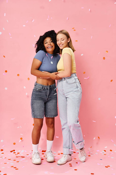 Two attractive diverse women in cozy casual attire standing next to each other on a pink background. - Photo, Image