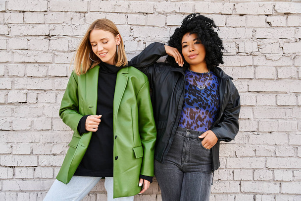 Dos mujeres de diferentes etnias en ropa casual se paran una al lado de la otra frente a una pared de ladrillo. - Foto, Imagen