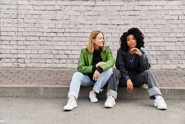 Deux femmes diverses en tenue décontractée assises sur un trottoir près d'un mur de briques. - Photo, image