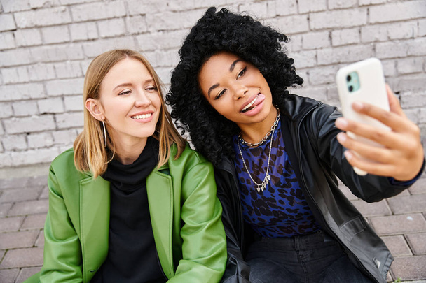 Dos mujeres en atuendo casual sentadas en el suelo, capturando un momento con una selfie. - Foto, imagen