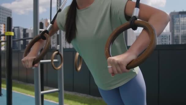 Inclinación lateral de la sonriente mujer caucásica usando anillos romanos para realizar flexiones en el campo deportivo de la azotea con hermoso paisaje urbano - Metraje, vídeo