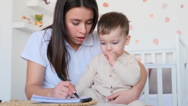Toddler and mother drawing or writing together in notebook, engaged in happy educational process, in playing room at home. Love between parent and child. - Footage, Video