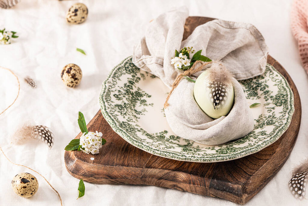 Stylish Easter brunch table setting with egg in easter bunny napkin. Modern natural dyed egg on napkin with bunny ears, flowers on vintage plate. Rustic Easter table decorations - 写真・画像